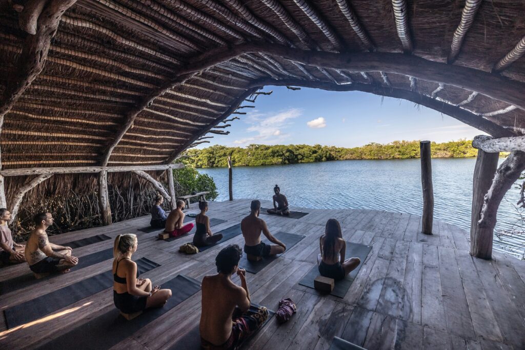 A group of people sitting on yoga mats under a large thatched-roof wooden structure overlooking a calm body of water.
